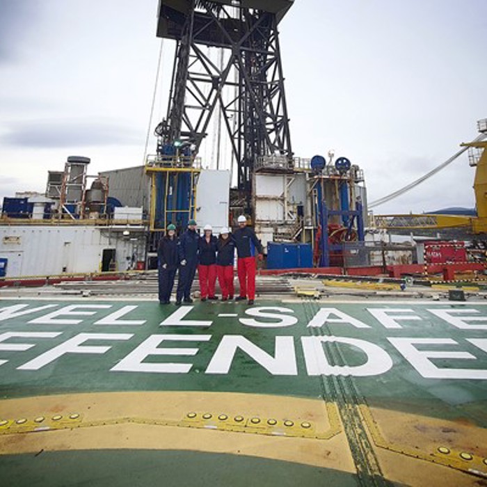 Quatro pessoas em uniformes de trabalho no convés da plataforma de perfuração offshore "Well-Safe Defender", com maquinaria industrial em fundo.