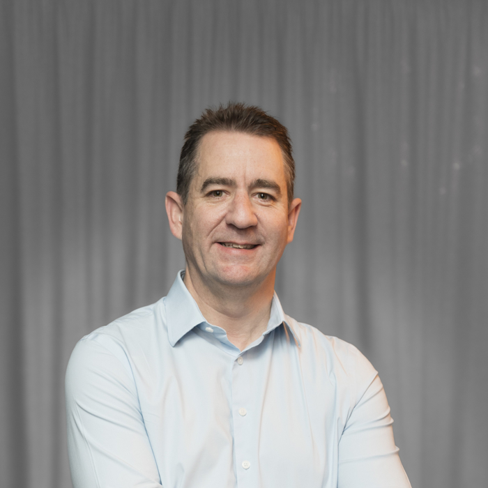 A man in a light blue shirt stands with his arms crossed, smiling, in front of a grey curtain background.