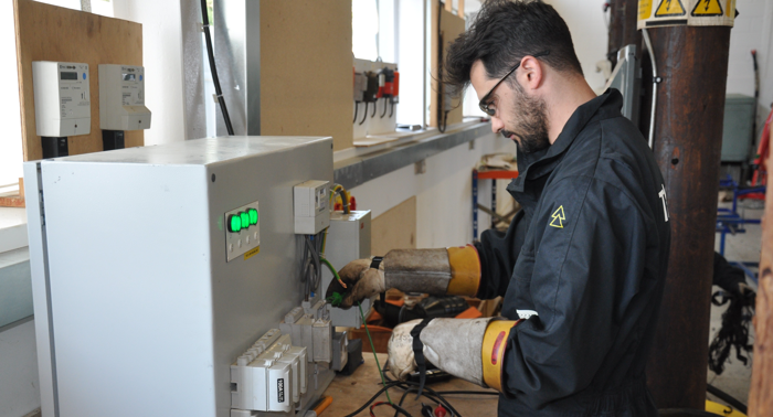A man wearing protective gear works on an electrical panel, adjusting wires with precision in a utilities and construction training workshop.