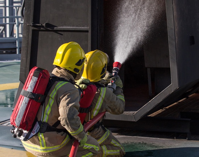 Firefighters in yellow helmets and protective gear spray water into a burning structure during a training exercise.