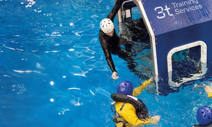 Two individuals in the water perform a safety exercise with one person reaching out from a cabin labeled "3t Training Services" while another person inside wears a helmet.