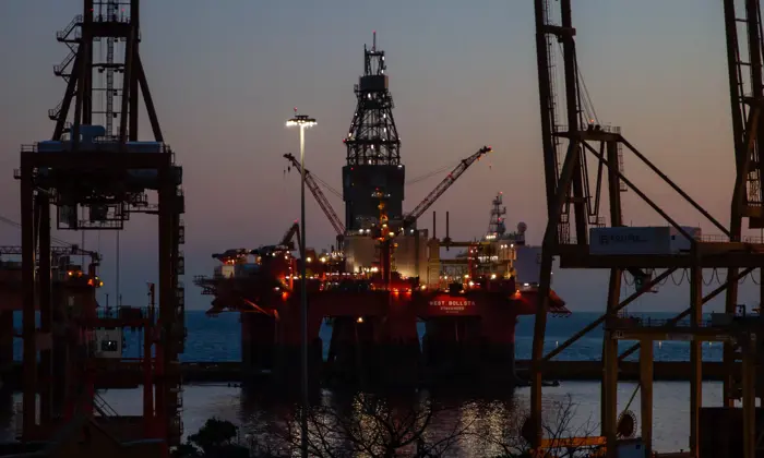 An offshore oil rig lit up at dusk, surrounded by the silhouettes of large cranes and industrial structures, with the sea in the background.