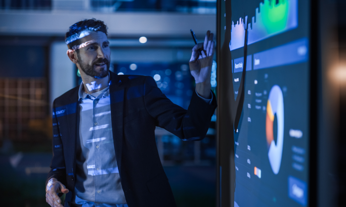 A man in a business suit points at a large interactive screen displaying graphs and charts in a modern office setting.