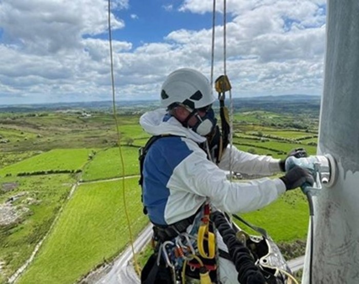 A person wearing safety gear and a helmet works on a structure high above a rural landscape with green fields under a partly cloudy sky.