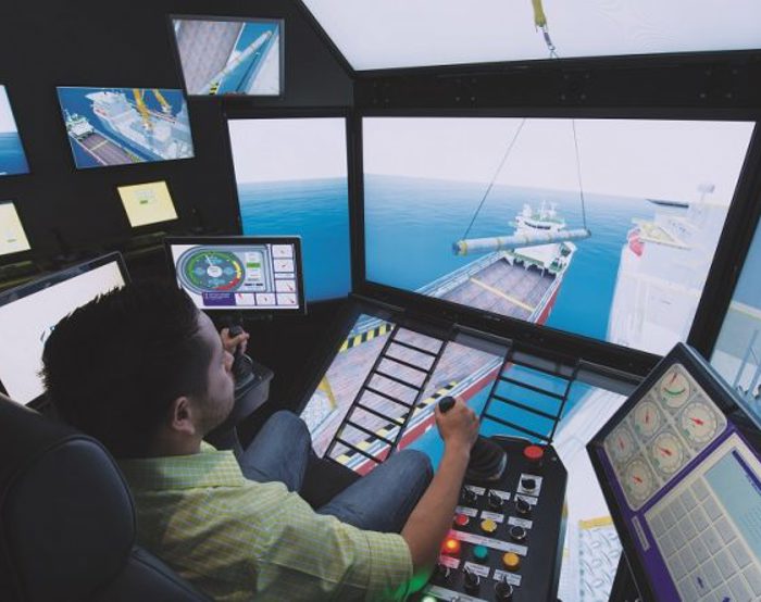 A person operates a complex maritime simulator with multiple screens, showing a ship being loaded. Various control panels and monitors are visible in the simulator room.