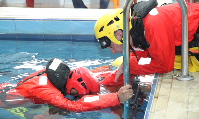 Two rescue workers in red suits and helmets perform a pool training exercise, with one standing in the water holding the pool edge and the other leaning over.