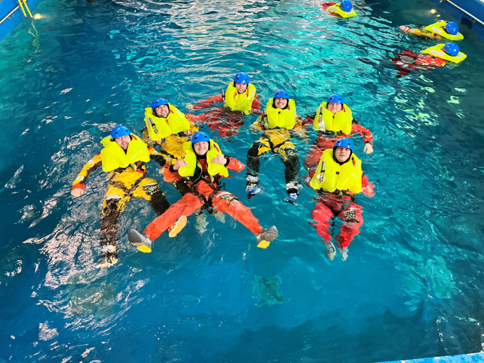 Seven individuals in bright yellow flotation suits and blue helmets float in a swimming pool, practicing water safety or rescue training techniques as precise as those performed by the military.