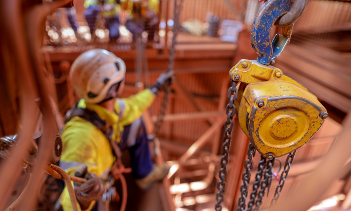 A construction worker in safety gear, undergoing training, is suspended by ropes next to a yellow hoist with chains.