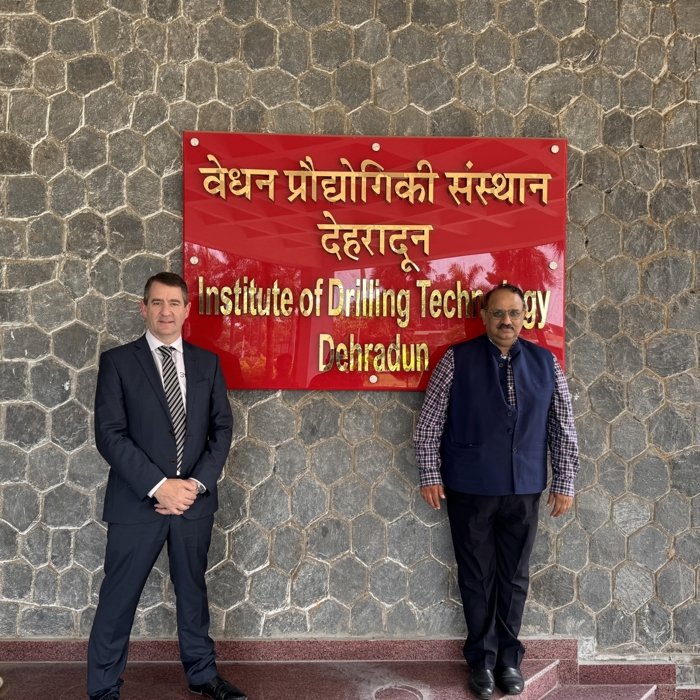 Deux personnes se tiennent devant un panneau rouge avec un texte en hindi et en anglais indiquant "Institute of Drilling Technology Dehradun", fixé à un mur de pierre.