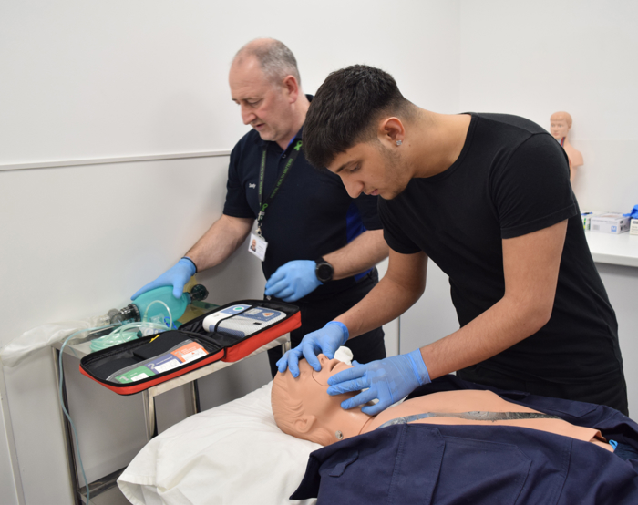 Two people practice CPR on a medical mannequin. One monitors equipment, while the other performs chest compressions. Both wear gloves.