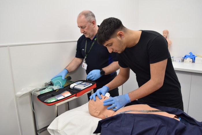 Two people practice CPR on a medical mannequin. One monitors equipment, while the other performs chest compressions. Both wear gloves.