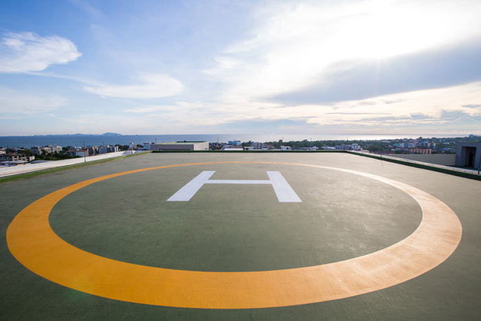 A rooftop helipad with a white "H" in the center is shown under a partly cloudy sky with a distant view of buildings and hills.