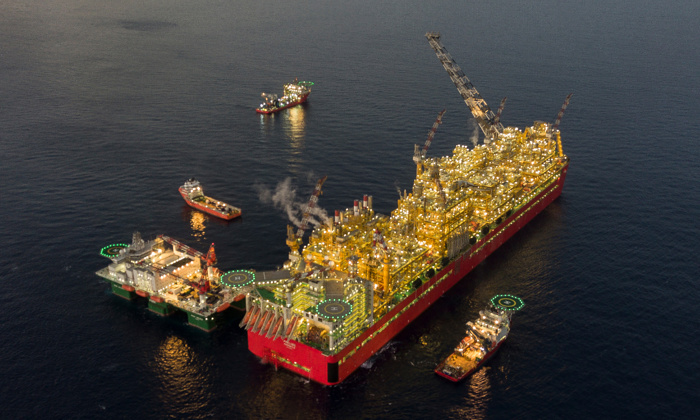 An offshore oil platform surrounded by support vessels floating in the sea. The platform is brightly lit and appears to be in operation, with smoke visible from its structures.