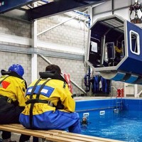 A group of people in yellow and blue safety gear sit on benches, observing a simulated helicopter cockpit suspended above a pool in an indoor training facility.