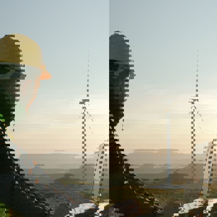 Person in a hard hat and safety vest using a laptop outdoors near wind turbines, with a scenic hilly landscape in the background during sunset.