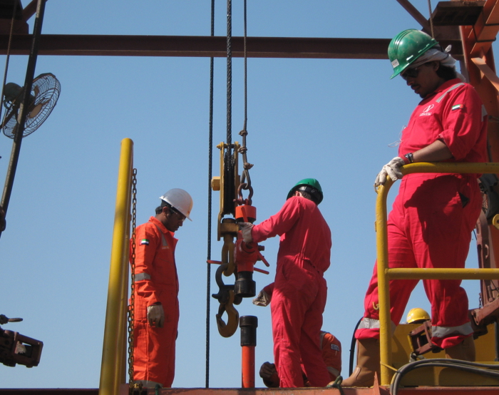 Workers in red coveralls and helmets operate equipment on an oil rig platform under a clear sky.