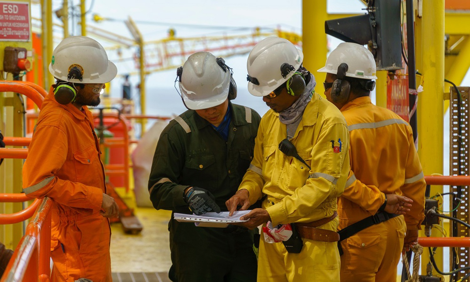 Four workers in safety gear review a document on an offshore oil rig. Two in orange coveralls, one in black, and one in yellow, discuss the contents while standing next to orange railings.
