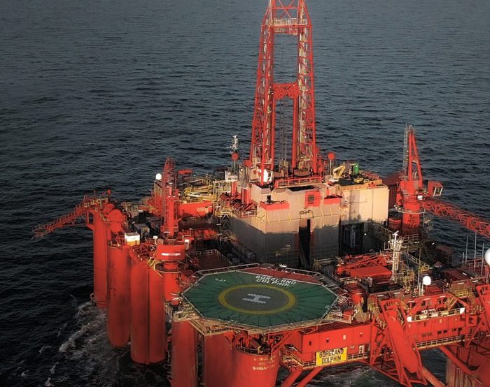 A red offshore oil drilling platform is positioned in open water, displaying a helipad and various equipment on its deck.