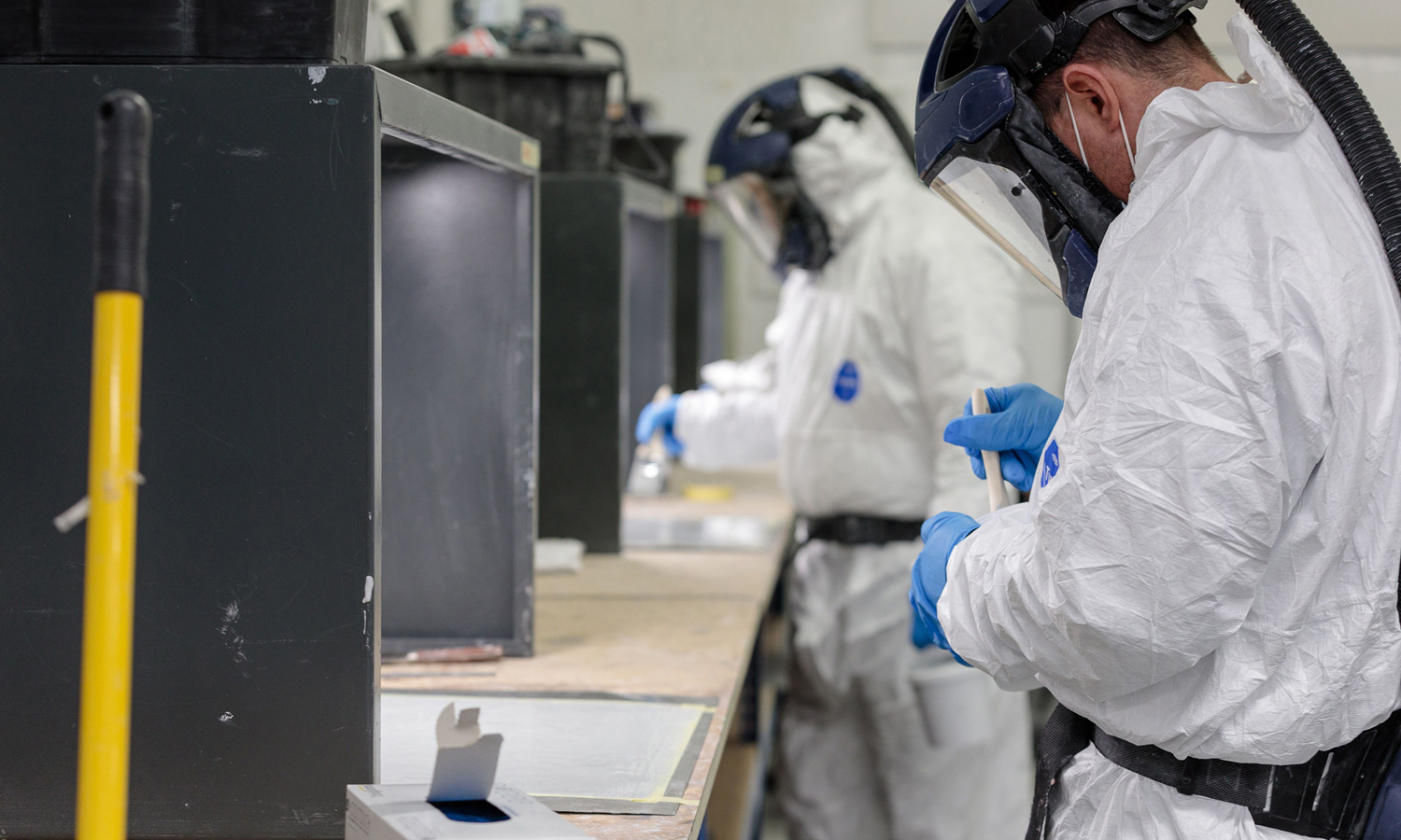 Two people in protective suits and helmets engage in a training session at the lab bench, carefully handling materials with gloves. A box and a yellow pole are visible in the foreground.