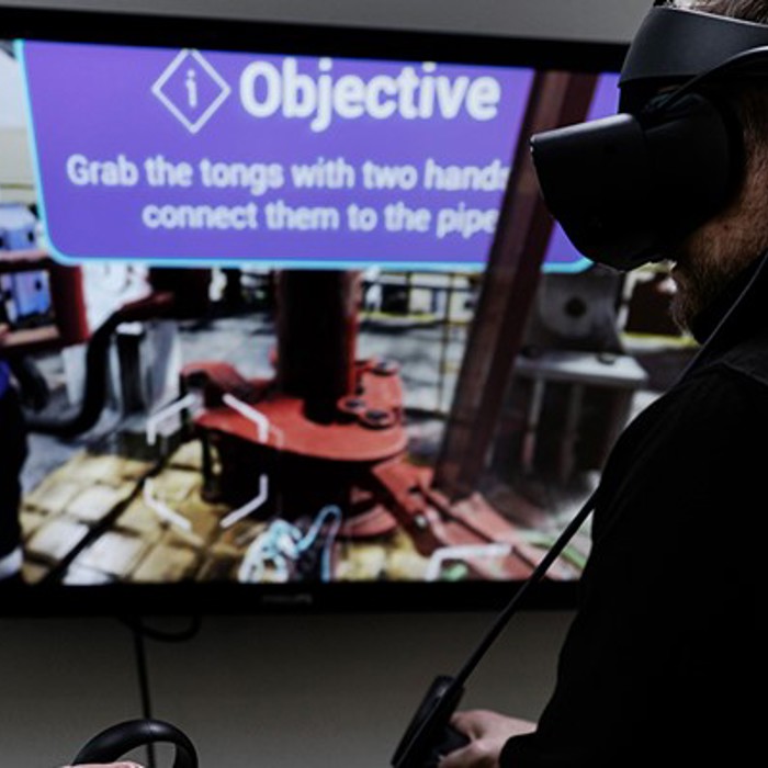 A person wearing a VR headset interacts with a virtual environment displayed on a screen, showing an instruction to "Grab the tongs with two hands and connect them to the pipe.