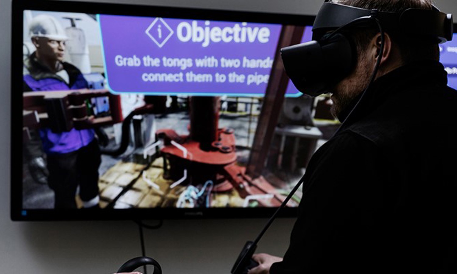 A person wearing a VR headset interacts with a virtual environment displayed on a screen, showing an instruction to "Grab the tongs with two hands and connect them to the pipe.