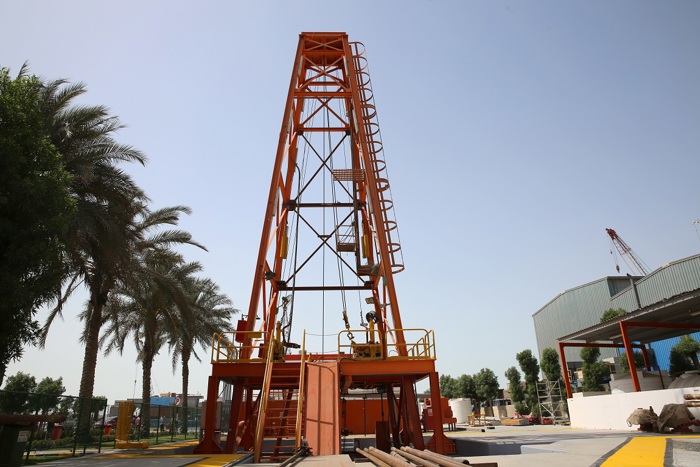 An orange drilling rig stands surrounded by trees with a clear sky in the background. Pipes are visible in the foreground.