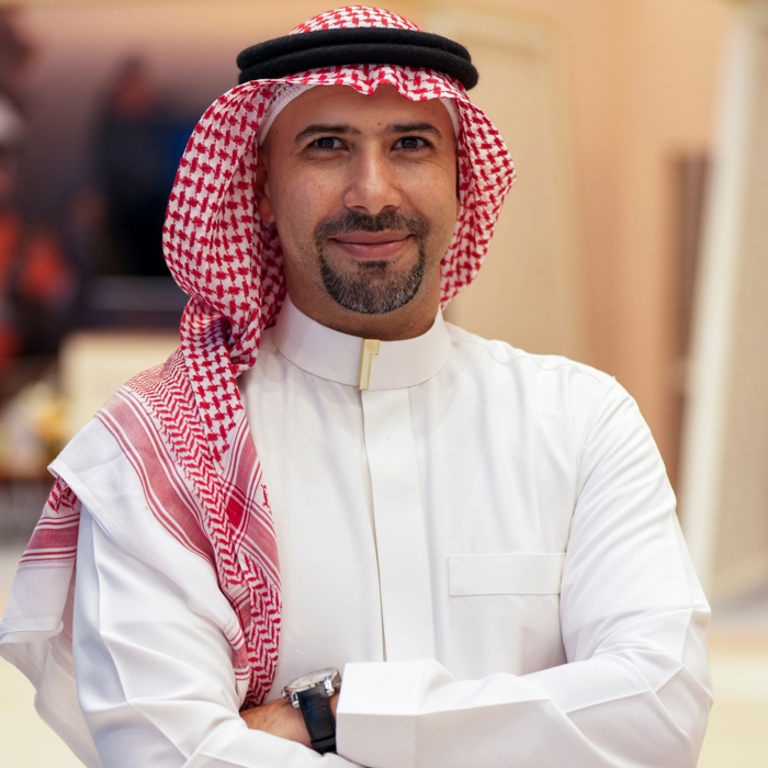 رجل ذو لحية يرتدي عباءة بيضاء وكوفية حمراء وبيضاء ويبتسم ويضع ذراعيه متشابكة.