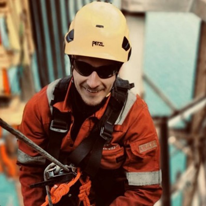 A worker wearing a yellow helmet and safety equipment is climbing a structure with ropes, with a large body of water visible in the background.