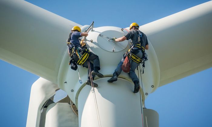 Two workers in safety harnesses and helmets are performing maintenance at the top of a wind turbine against a clear blue sky.