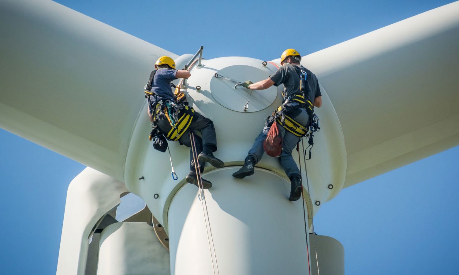 Two workers in safety harnesses and helmets are performing maintenance at the top of a wind turbine against a clear blue sky.