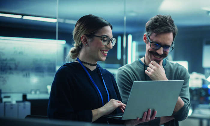 Two people are standing and collaborating in an office, looking at a laptop screen together and smiling. The room has a whiteboard and modern lighting.