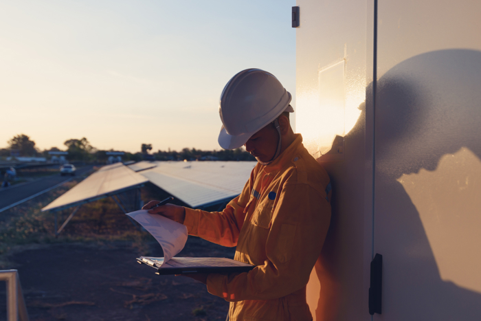 A worker in a yellow uniform and hard hat reads documents outside at sunset, leaning against a wall next to solar panels.
