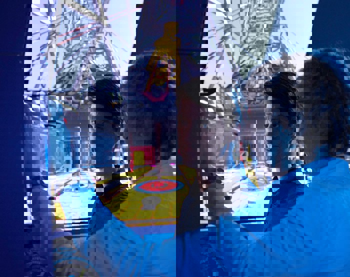 Two men in a control room operate a simulator with a large screen displaying the interior of an industrial setting. One is pointing while the other observes intently.