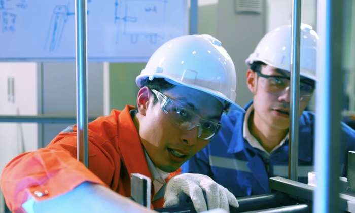 Two workers in hard hats and safety glasses are examining machinery in a facility. One worker in an orange jumpsuit is pointing, while the other in a blue jumpsuit observes. A whiteboard is visible behind them.