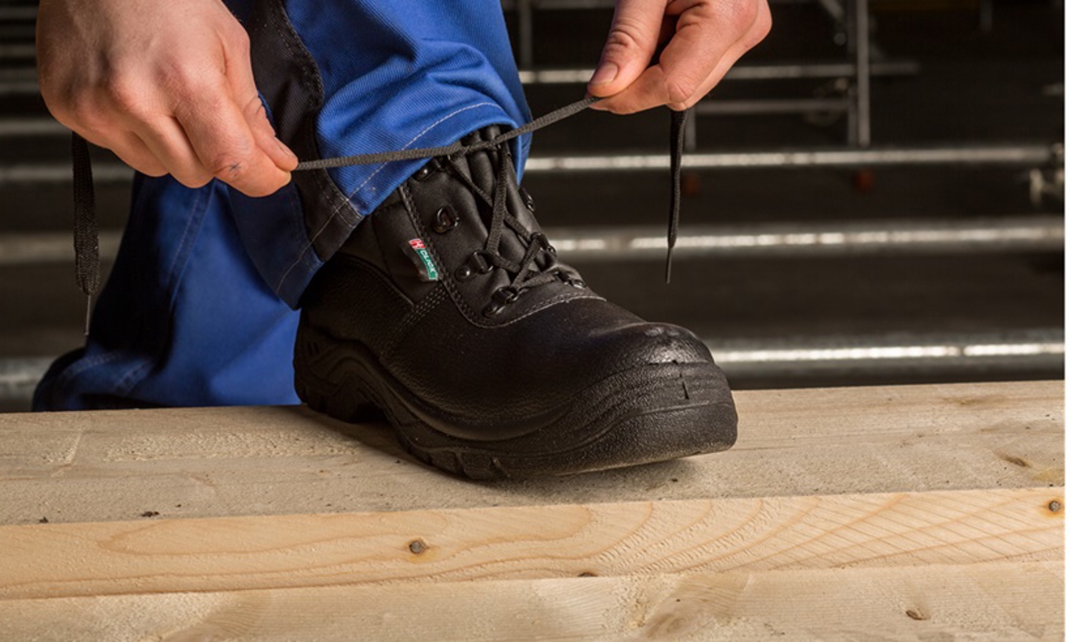 Standing on a wooden surface, the person focuses intently, as if in training, tying the laces of a sturdy black work boot.