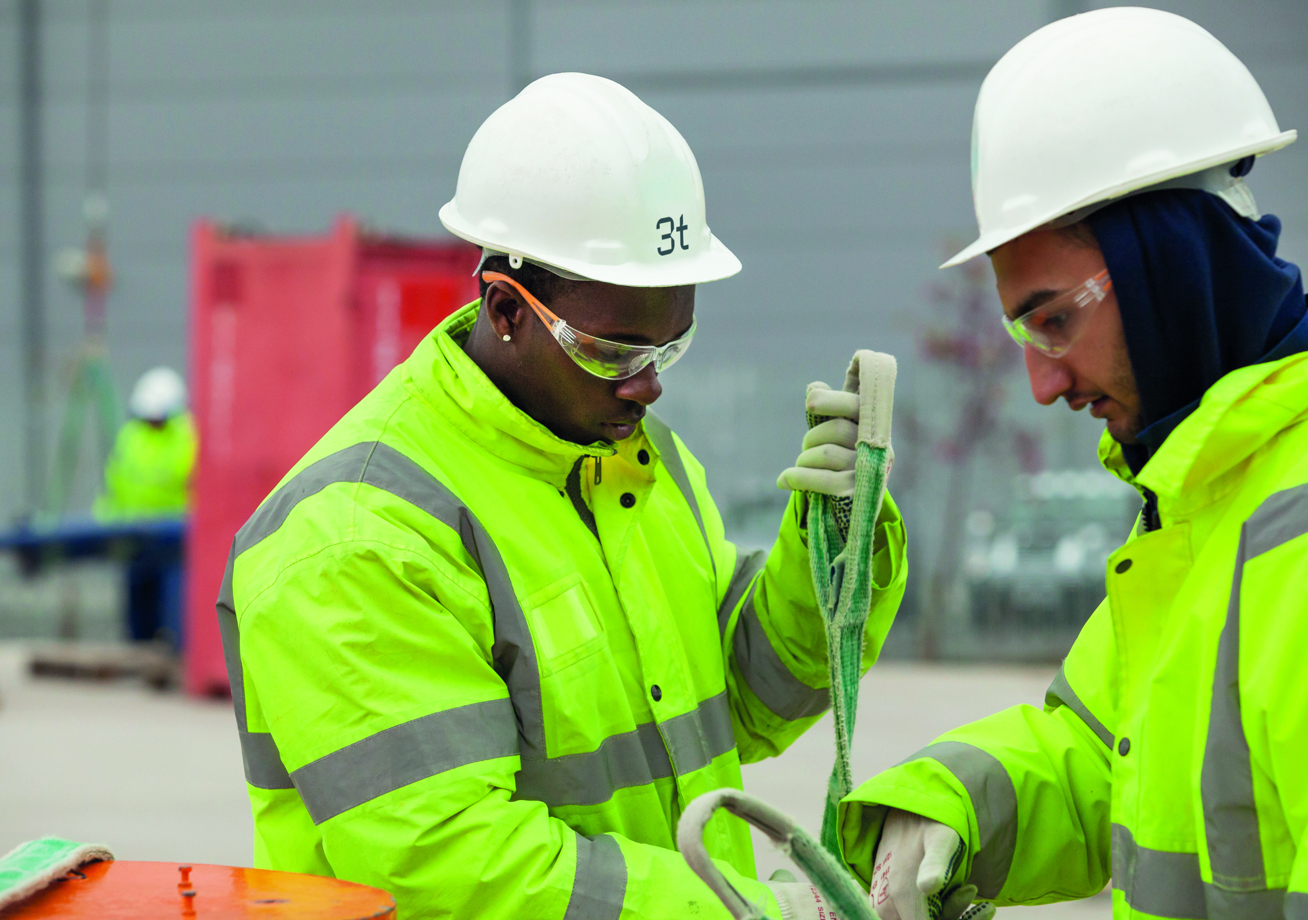 Two workers wearing high-visibility jackets and safety helmets are undergoing training as they skillfully handle a rope or strap outdoors.